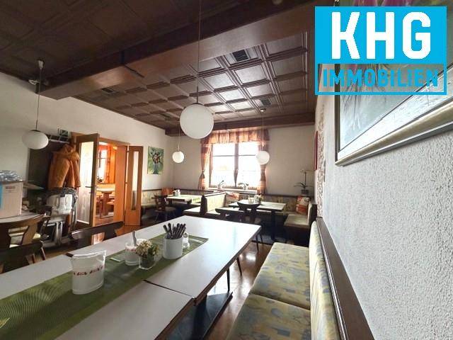 Weiterer Gastraum mit Holzbalkendecke, schlichten Tischen und Bänken sowie einem Fenster mit Vorhängen.