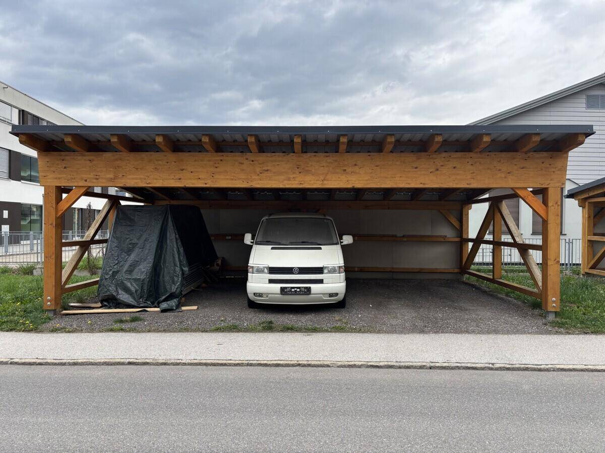 Geräumiger Holzcarport mit einem weißen Lieferwagen darunter, auf einem Schottergrund.