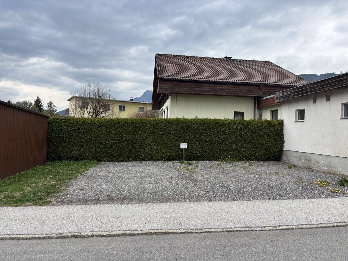 Schotterparkplatz mit einer hohen Hecke im Hintergrund und einem Wohnhaus.
