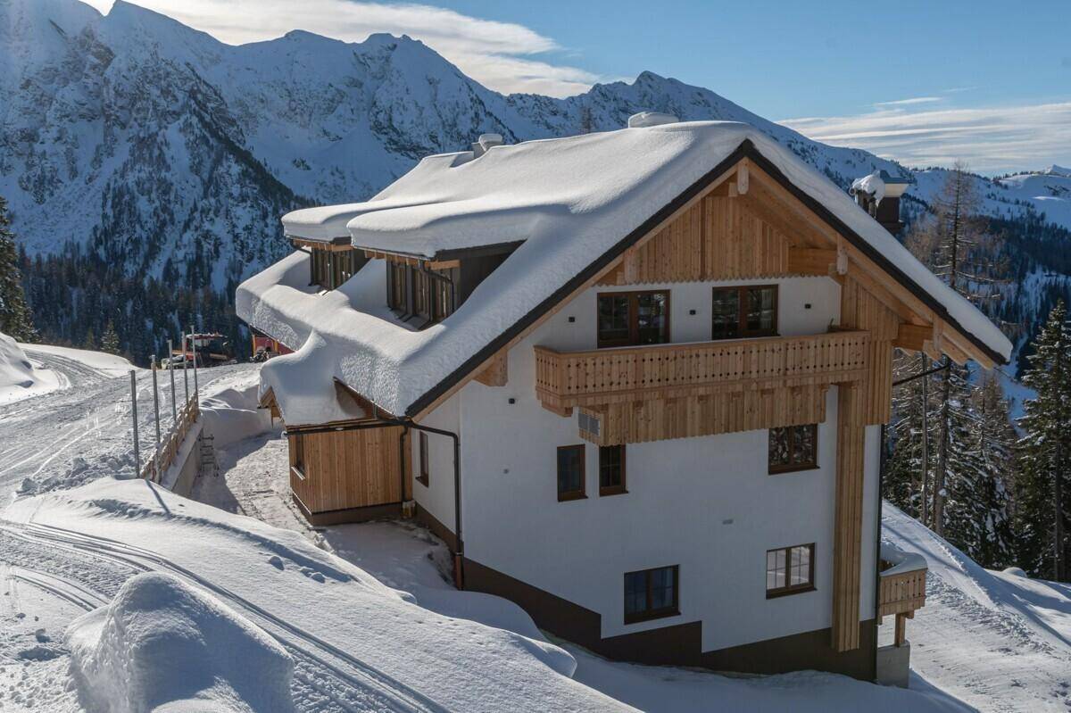 Alpenhaus mit Holzbalkonen und schneebedecktem Dach, umgeben von verschneiten Bergen und Wäldern.