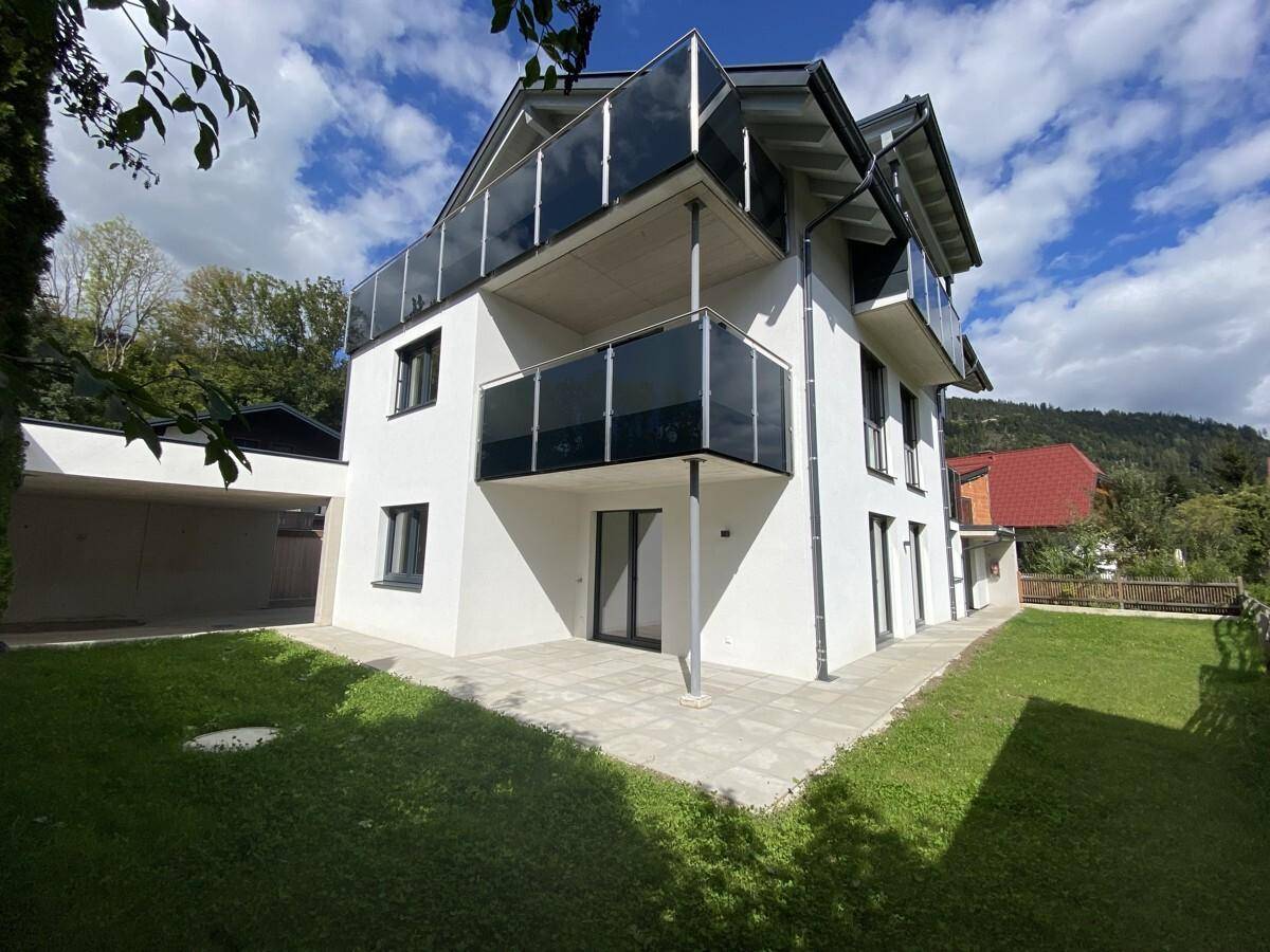 Neubau mit Terrasse und Gartenbereich, umgeben von grüner Landschaft.