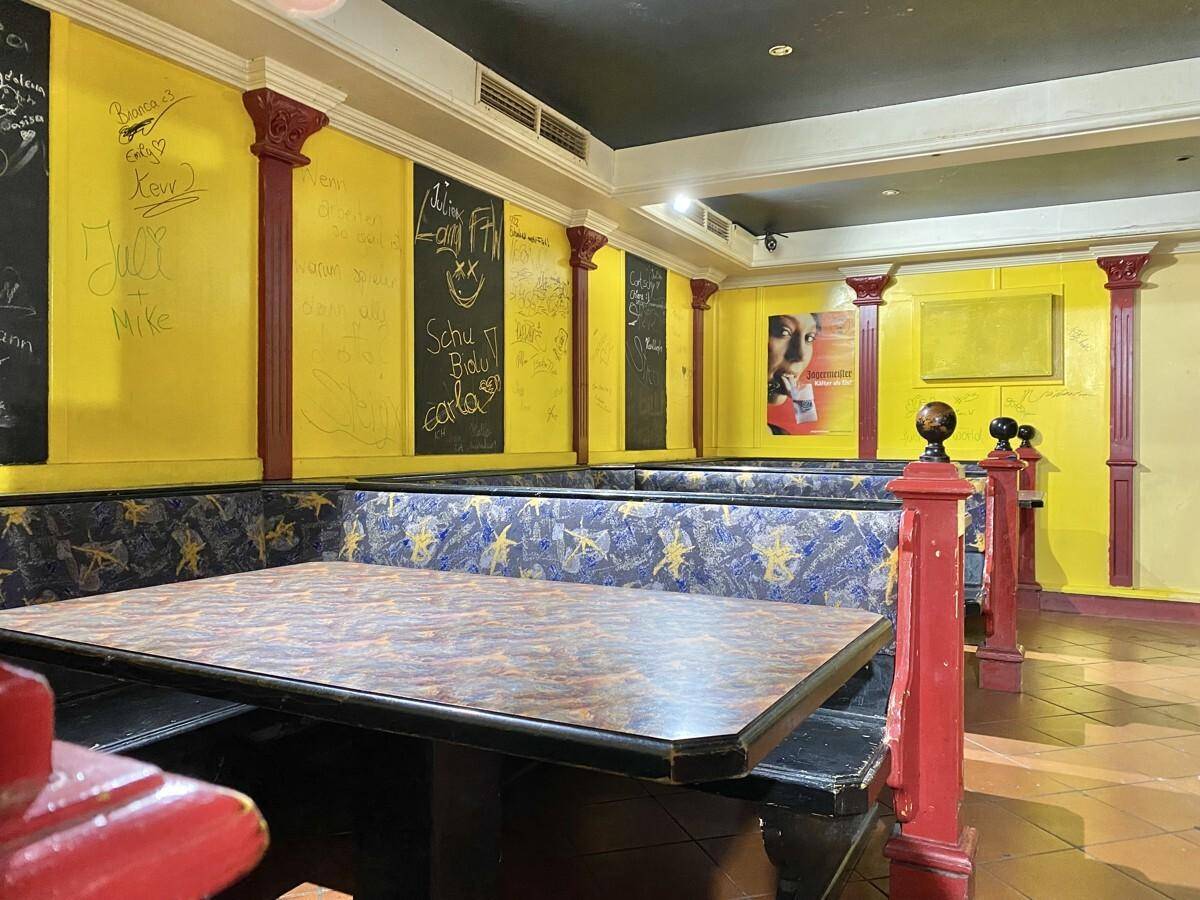 Sitzbereich einer Bar mit gepolsterten Bänken, Tischen und gelben Wänden, verziert mit roten Säulen und Graffiti.