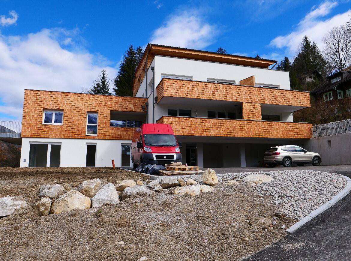Neubau mit Holzschindelfassade und Parkplätzen in einer bergigen Landschaft.