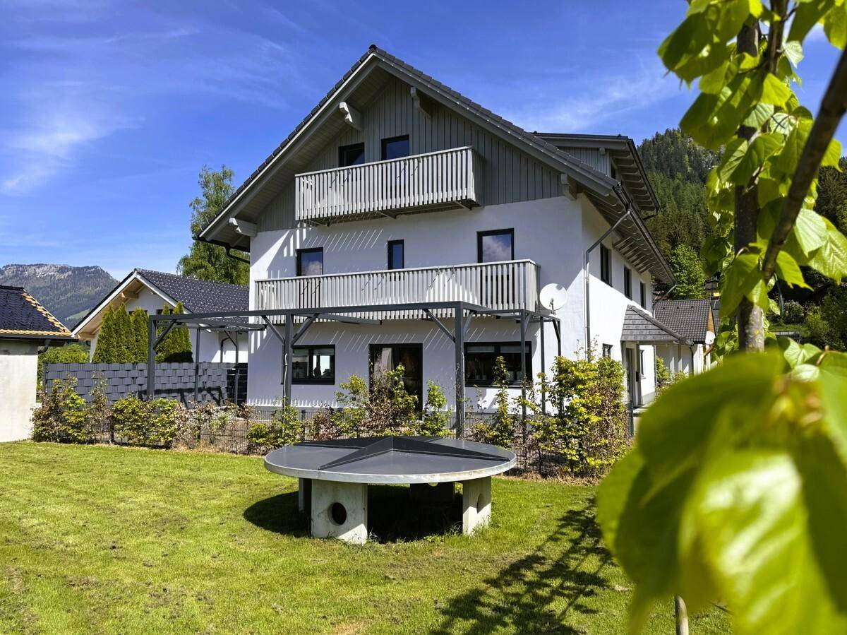 Gepflegter Garten mit Sitzgelegenheit und Blick auf das moderne Haus mit Balkonen und Bergen.