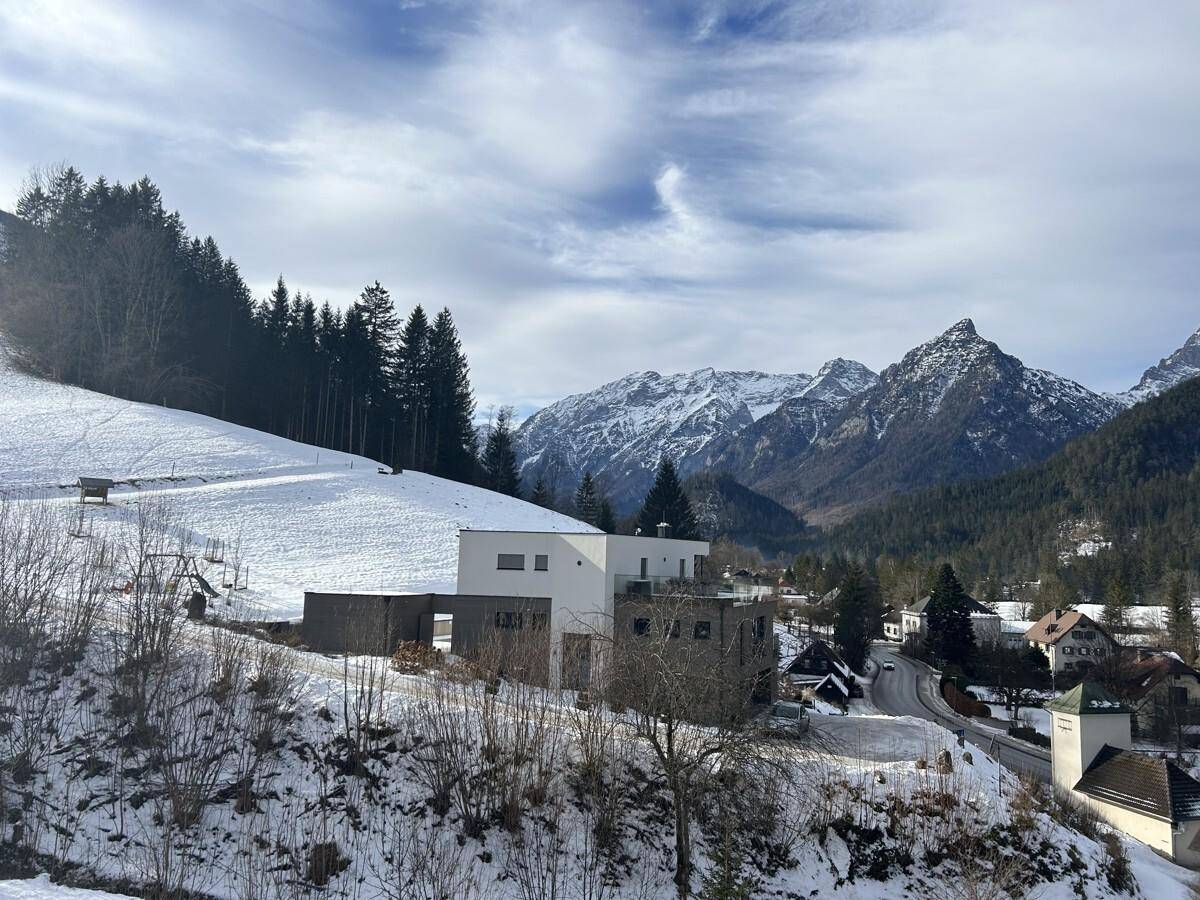 Weitläufiger Blick über ein schneebedecktes Tal mit einem modernen Gebäude und fernen Berggipfeln.
