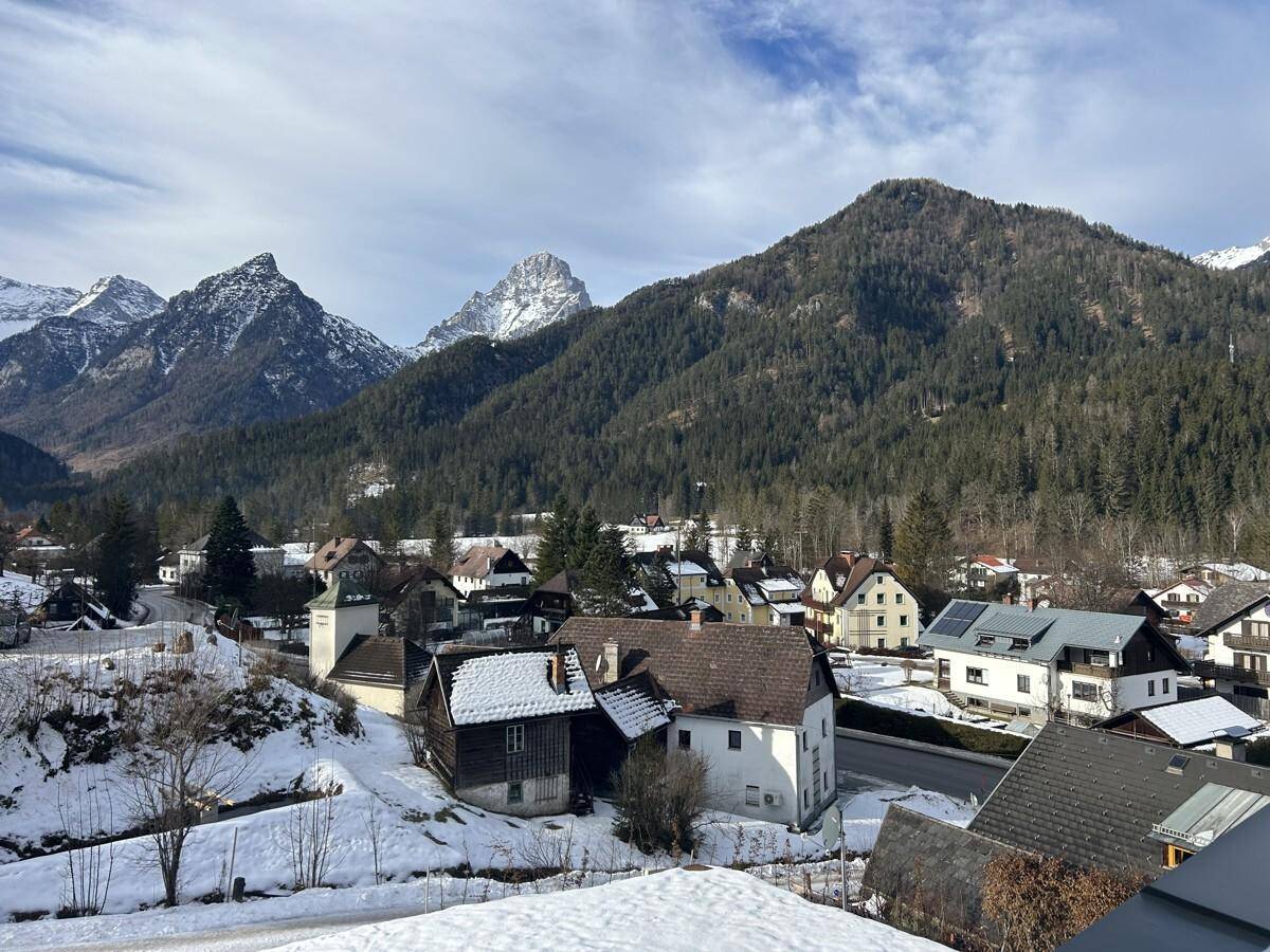 Malerischer Blick auf ein verschneites Dorf in einem Tal, umgeben von bewaldeten Bergen unter blauem Himmel.