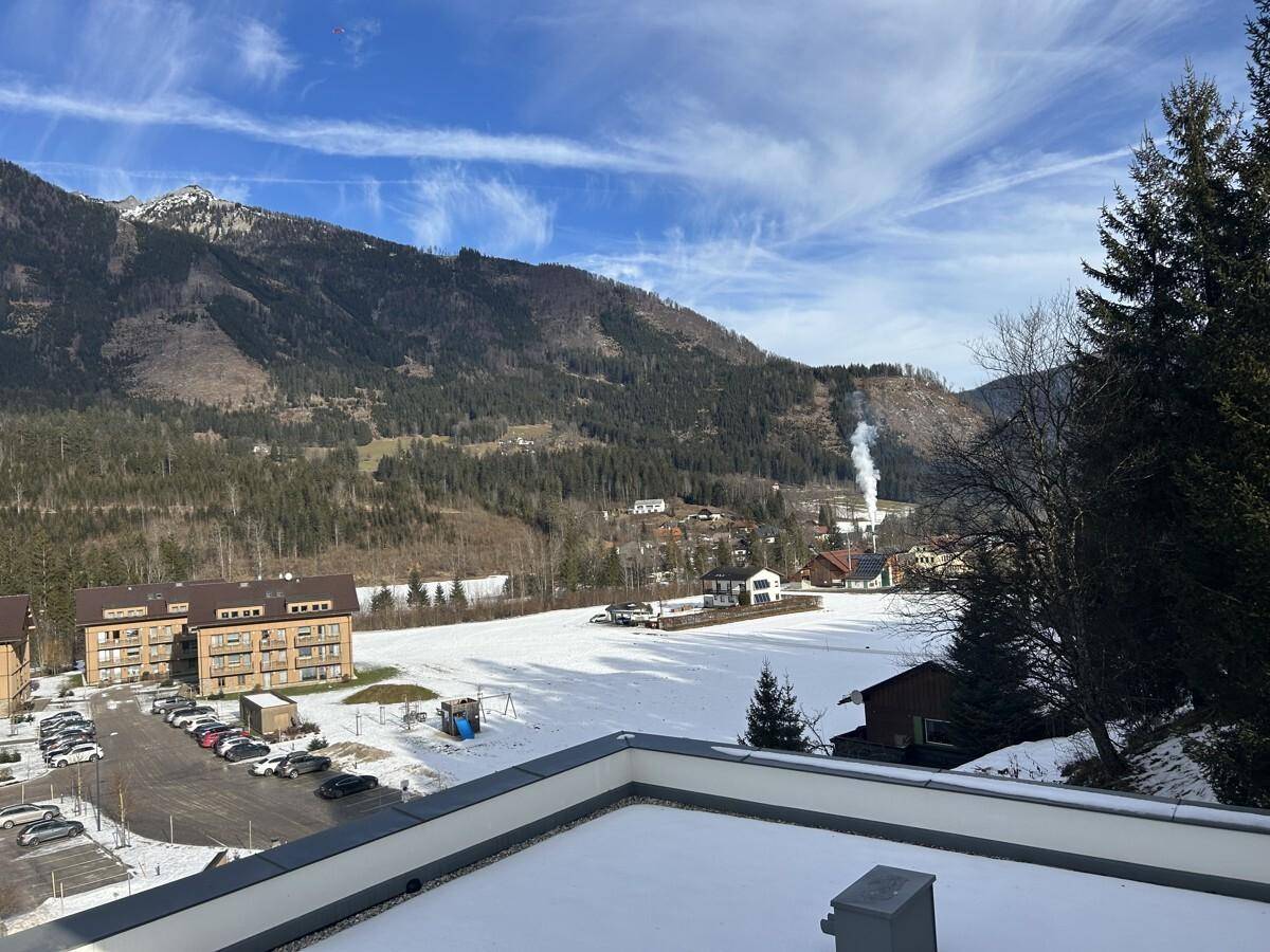 Blick von einer verschneiten Dachterrasse auf ein schneebedecktes Feld, Parkplätze und Berge.