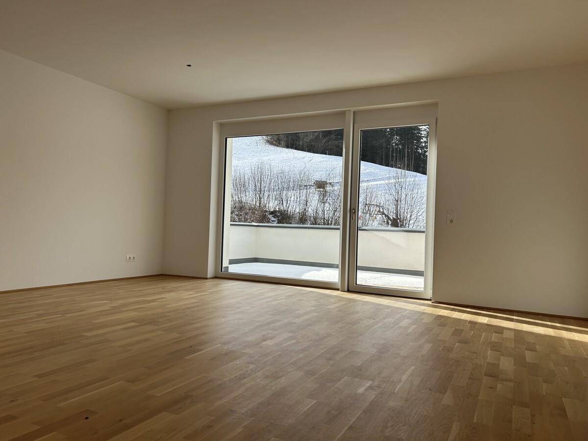 Helles, leeres Wohnzimmer mit Holzboden und großen Schiebetüren, die zu einem Balkon führen.