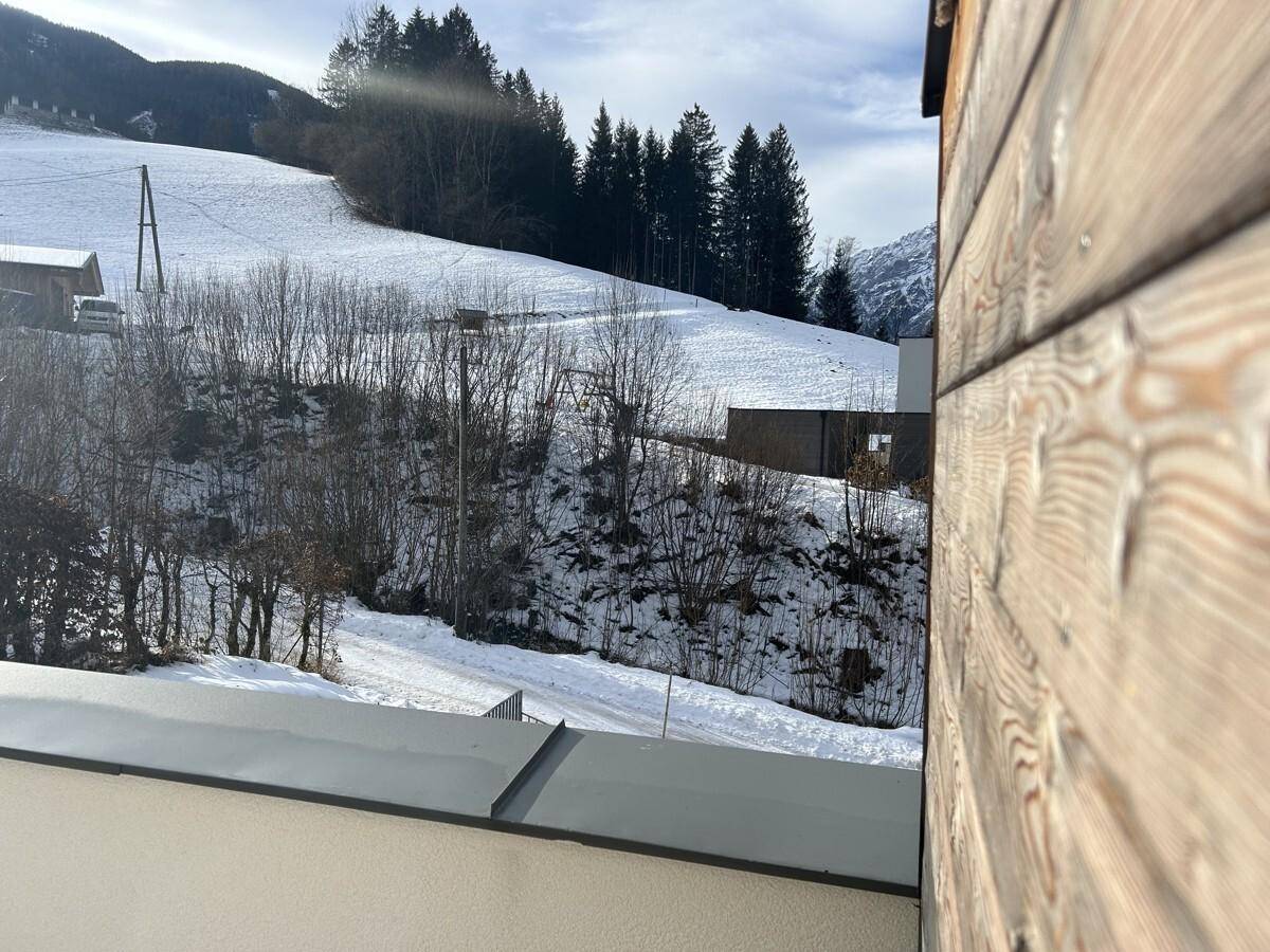 Blick auf eine schneebedeckte Hügellandschaft mit Bäumen, teilweise verdeckt von einer Holzfassade.
