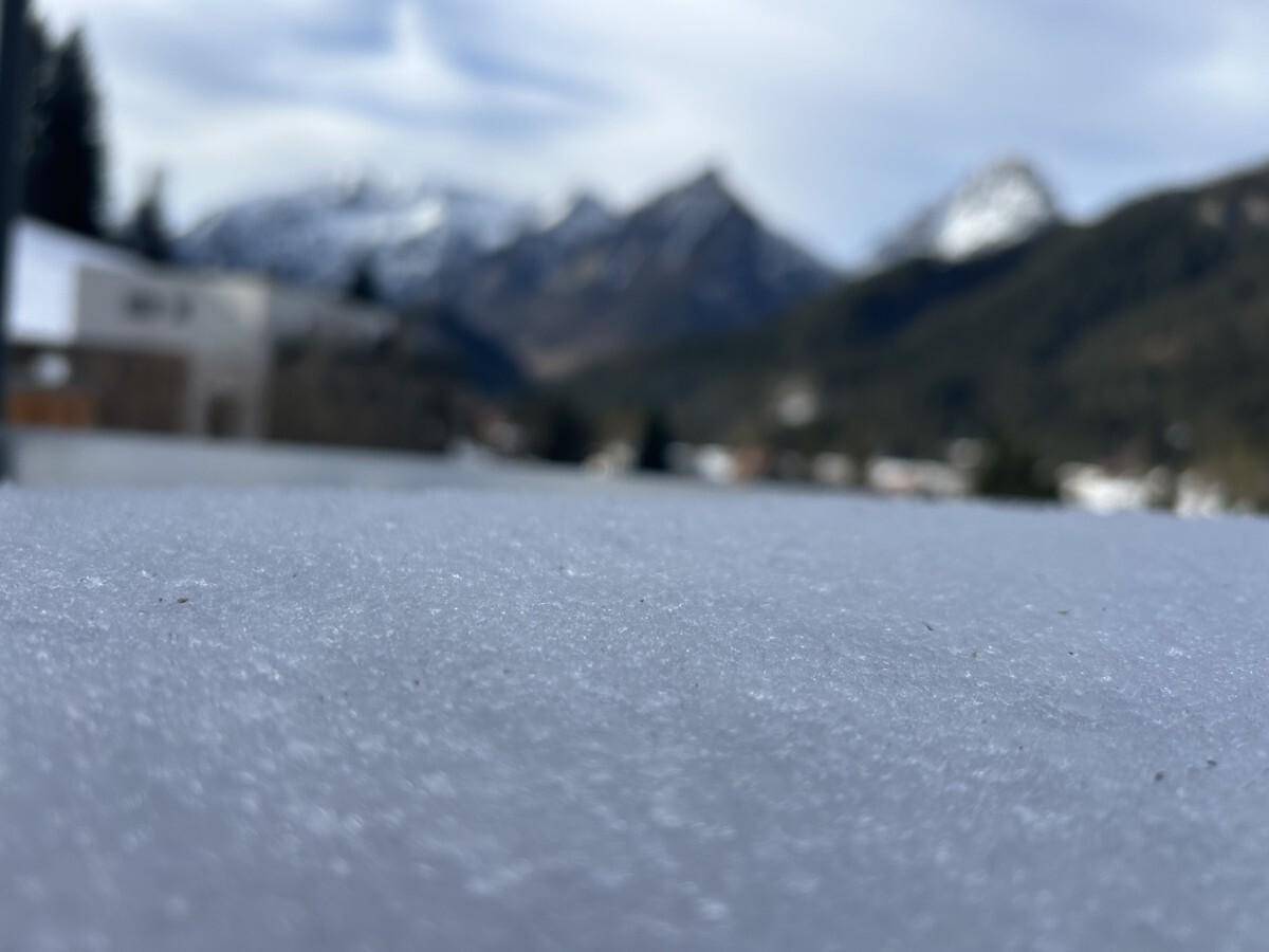 Detailansicht von frischem Schnee auf einer Terrasse mit verschwommenem Panorama der Alpen.