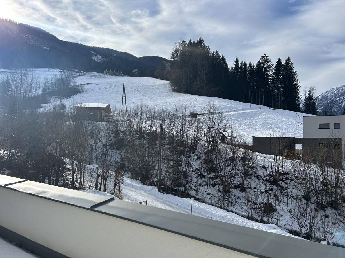 Weitläufiger Blick auf eine schneebedeckte Hügellandschaft mit Wald und einem kleinen Gebäude.