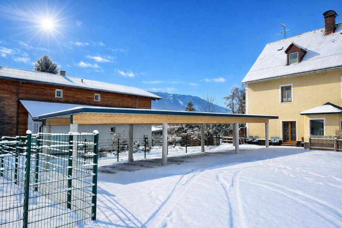 Carport mit Holzbalken und Betonsäulen, umgeben von Schnee und einem traditionellen Gebäude.
