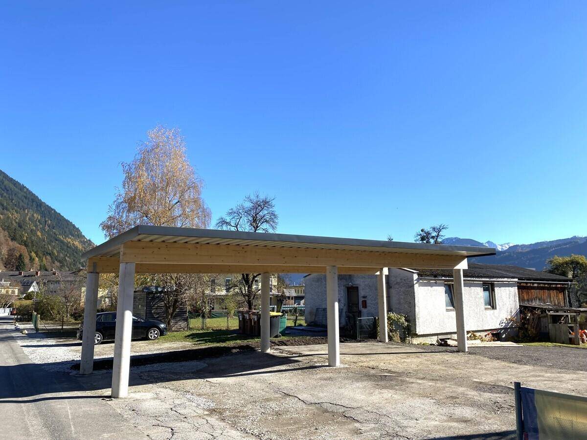 Offener Carport mit Holzdach und stabilen Säulen, umgeben von ländlicher Landschaft und Bergen.