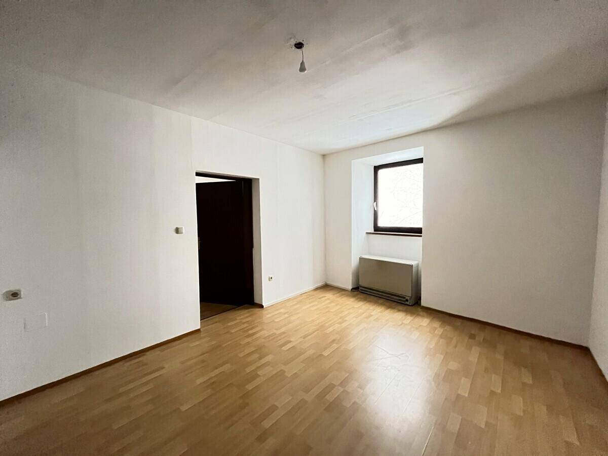 Geräumiges Wohnzimmer mit hellem Parkettboden und einem Fenster, das Tageslicht hereinlässt.