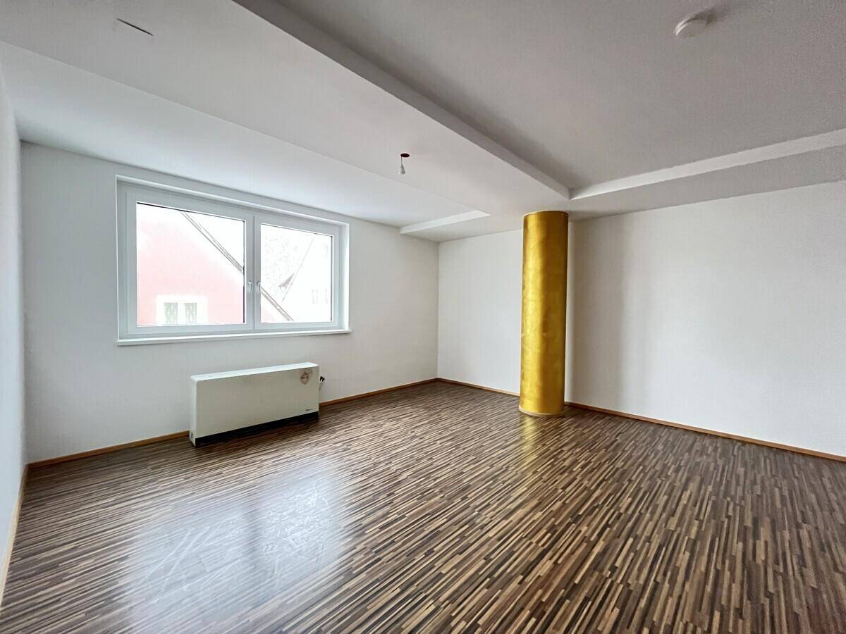 Leerer Raum mit dunklem Holzboden, weiß gestrichenen Wänden und einer goldenen Säule.