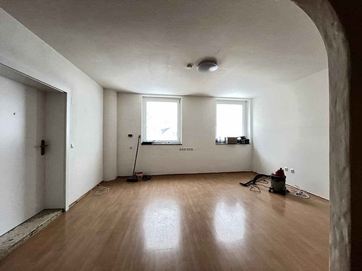 Geräumiges, leeres Wohnzimmer mit zwei Fenstern und hellem Parkettboden.
