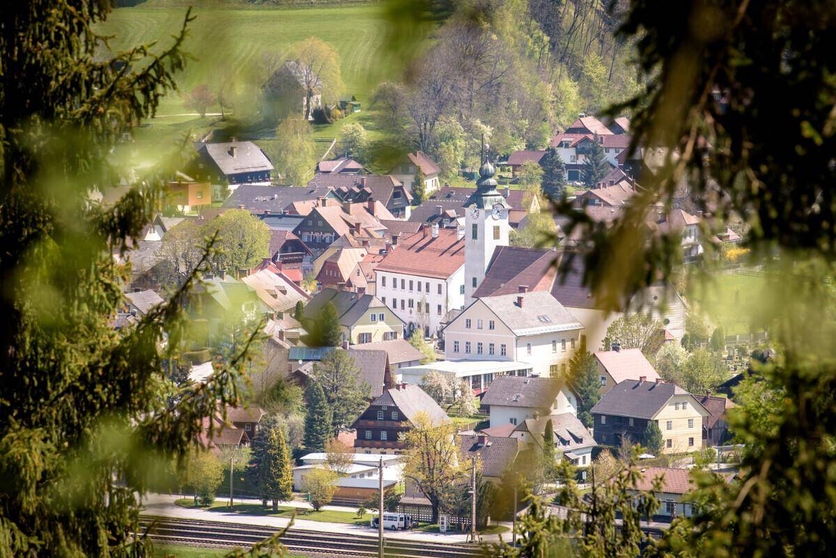 Blick durch Baumäste auf ein idyllisches Dorf mit Kirche und traditionellen Häusern.