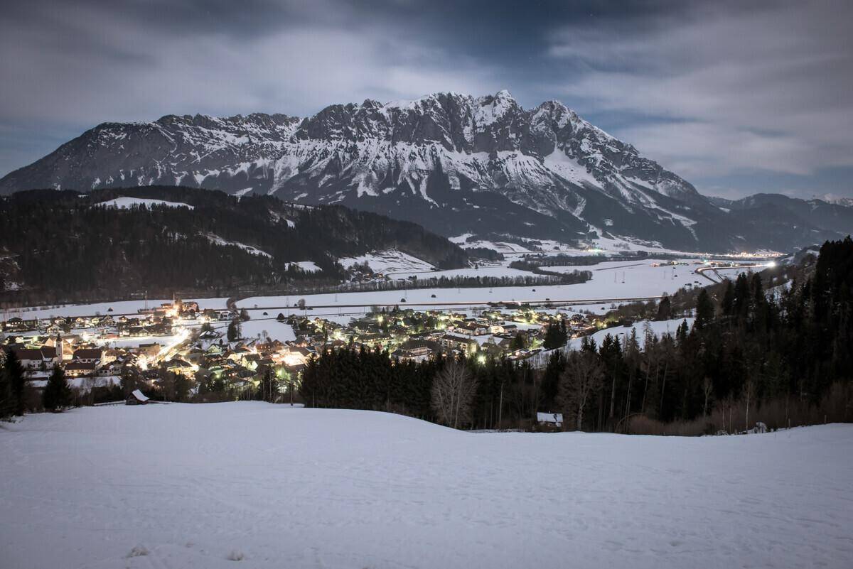 Winterliche Nachtansicht eines beleuchteten Dorfes in einem verschneiten Tal mit imposanten Bergen.