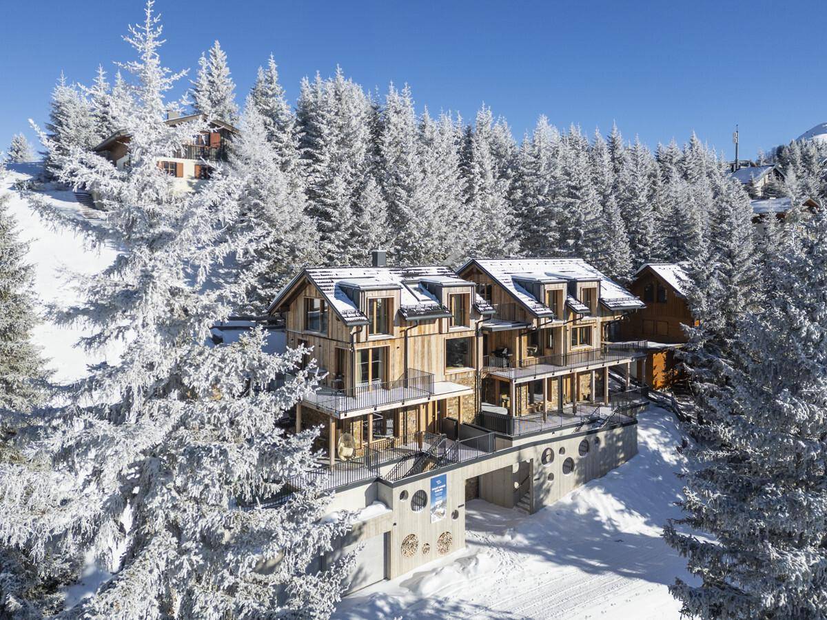 Luftaufnahme des modernen Holzgebäudes in einer weiten, verschneiten Berglandschaft mit Wald.