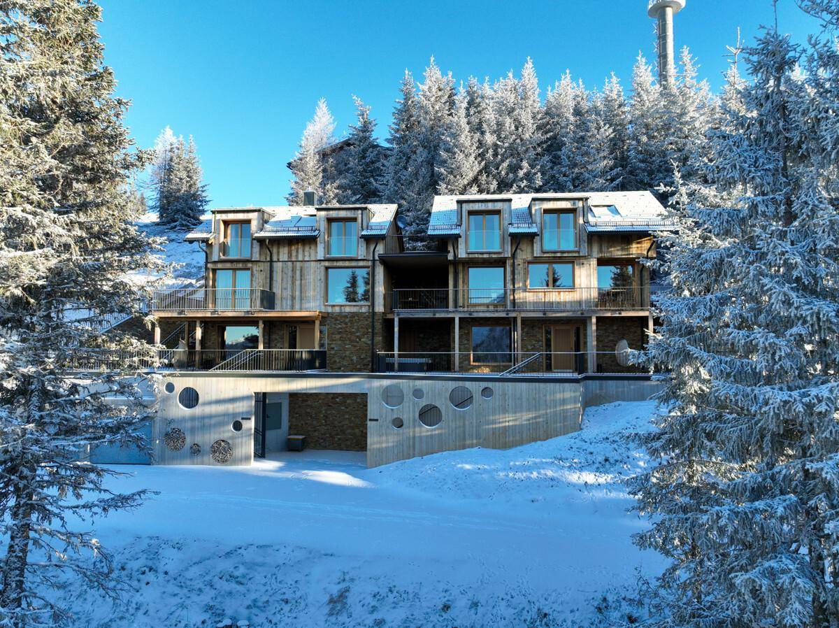 Modernes Holzgebäude in einer verschneiten, bewaldeten Berglandschaft unter blauem Himmel.