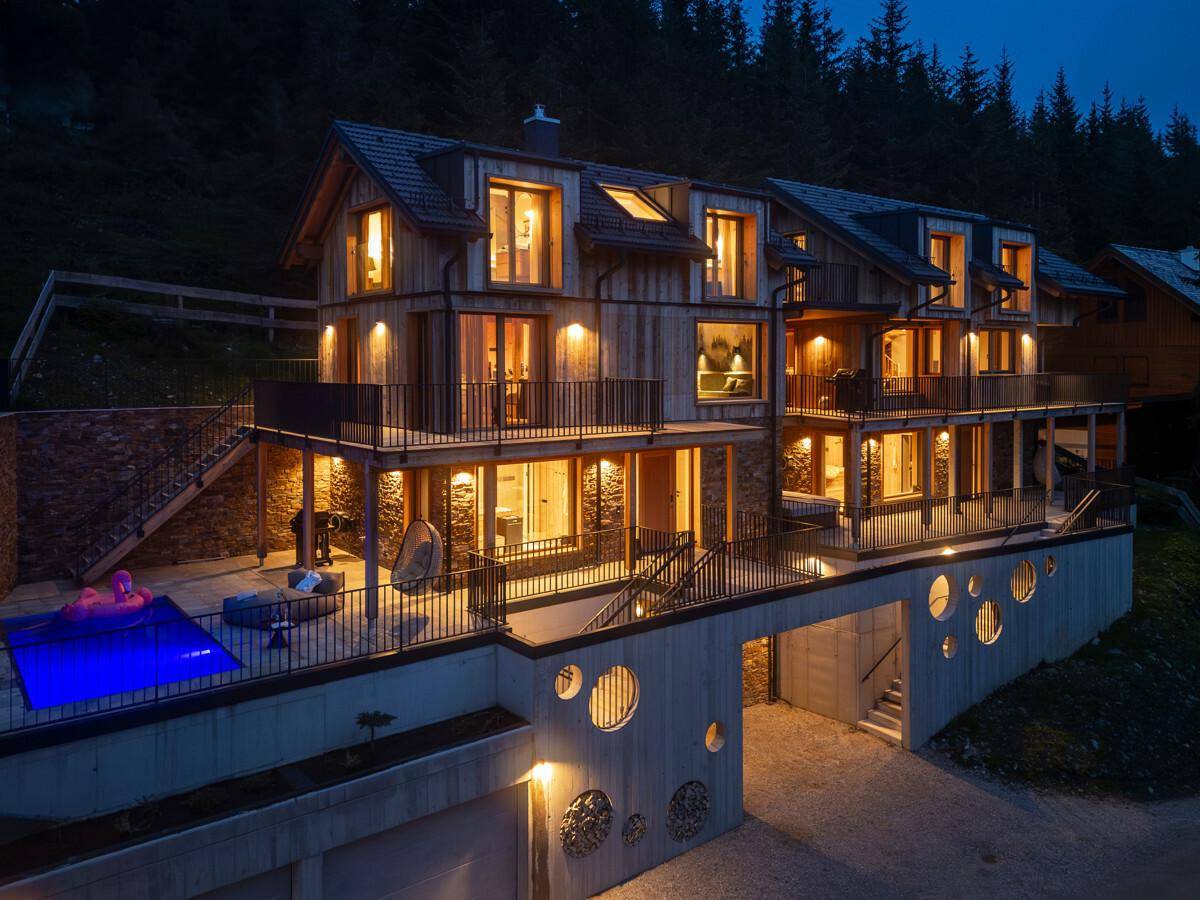 Modernes Holzgebäude bei Nacht mit beleuchteten Innenräumen, Balkonen und einem Außenpool.