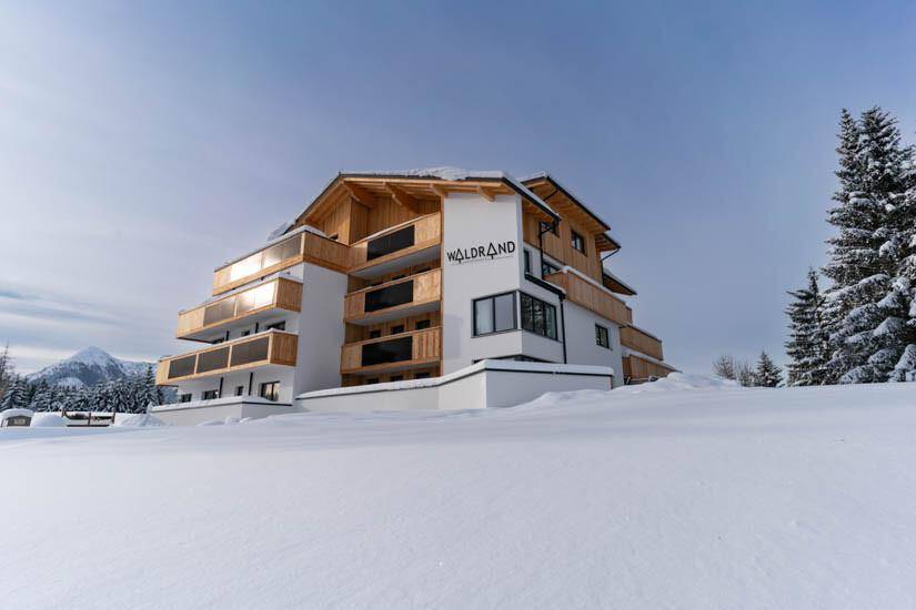 Modernes Gebäude in verschneiter Winterlandschaft, umgeben von Bäumen und Bergen.