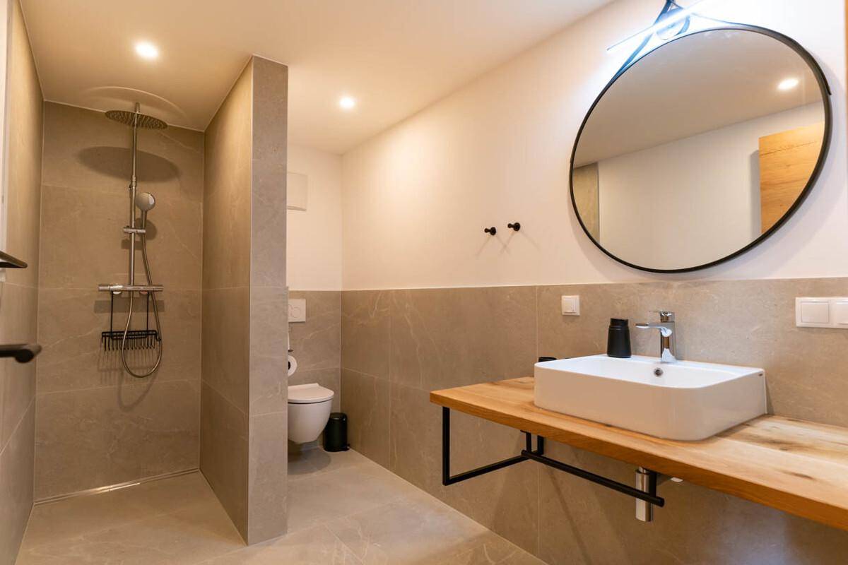 Modernes Badezimmer mit ebenerdiger Dusche, Waschtisch aus Holz und rundem Spiegel.