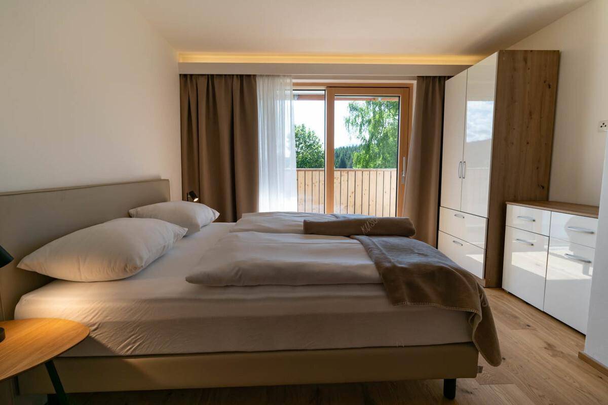 Helles Schlafzimmer mit Doppelbett, Holzboden und Zugang zu einem Balkon mit Grünblick.