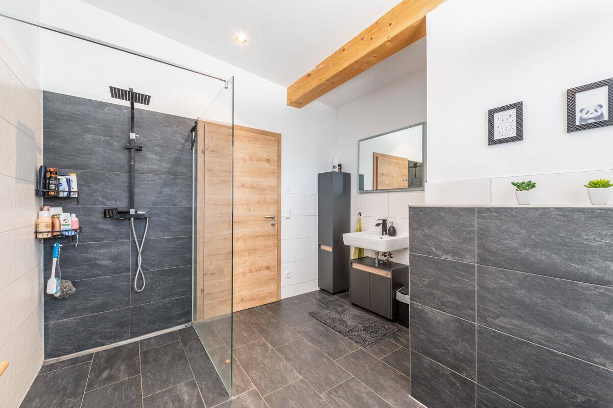 Modernes Badezimmer mit ebenerdiger Dusche, dunklen Fliesen und sichtbarem Holzbalken.