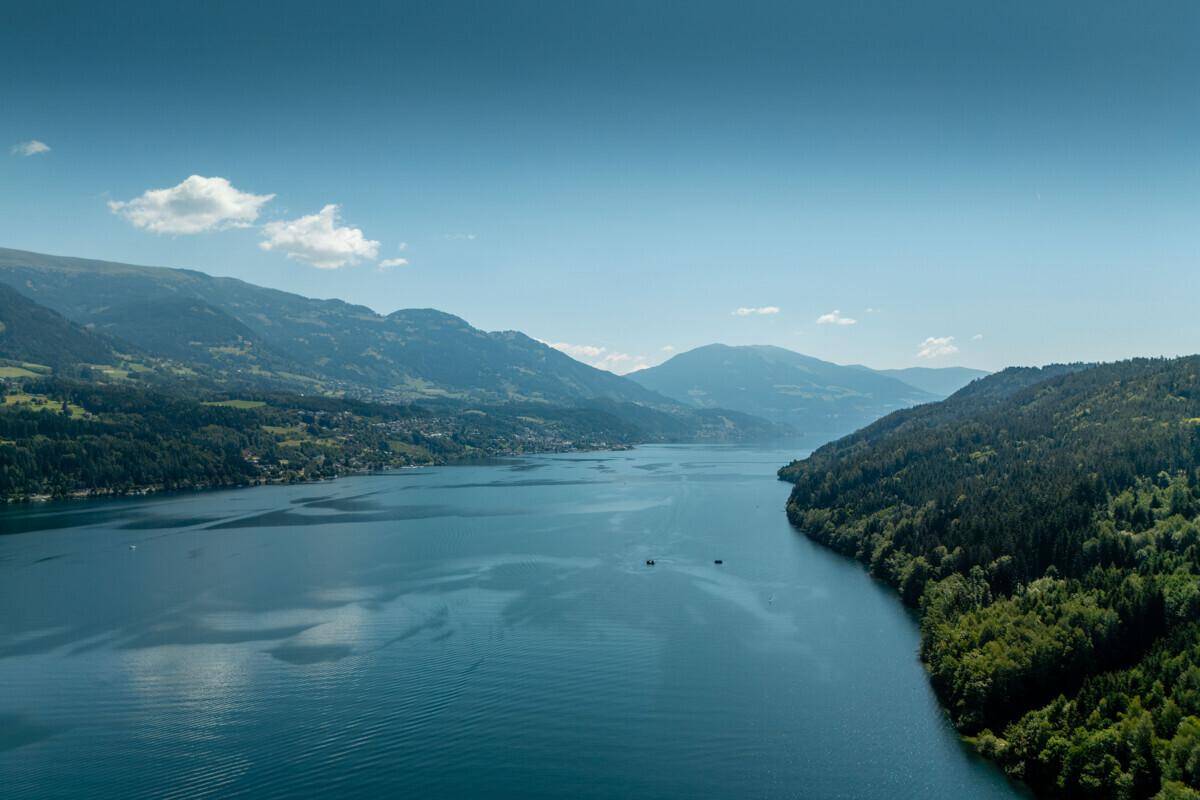 Weitläufiger Blick über den See und die bewaldeten Hügel unter blauem Himmel.