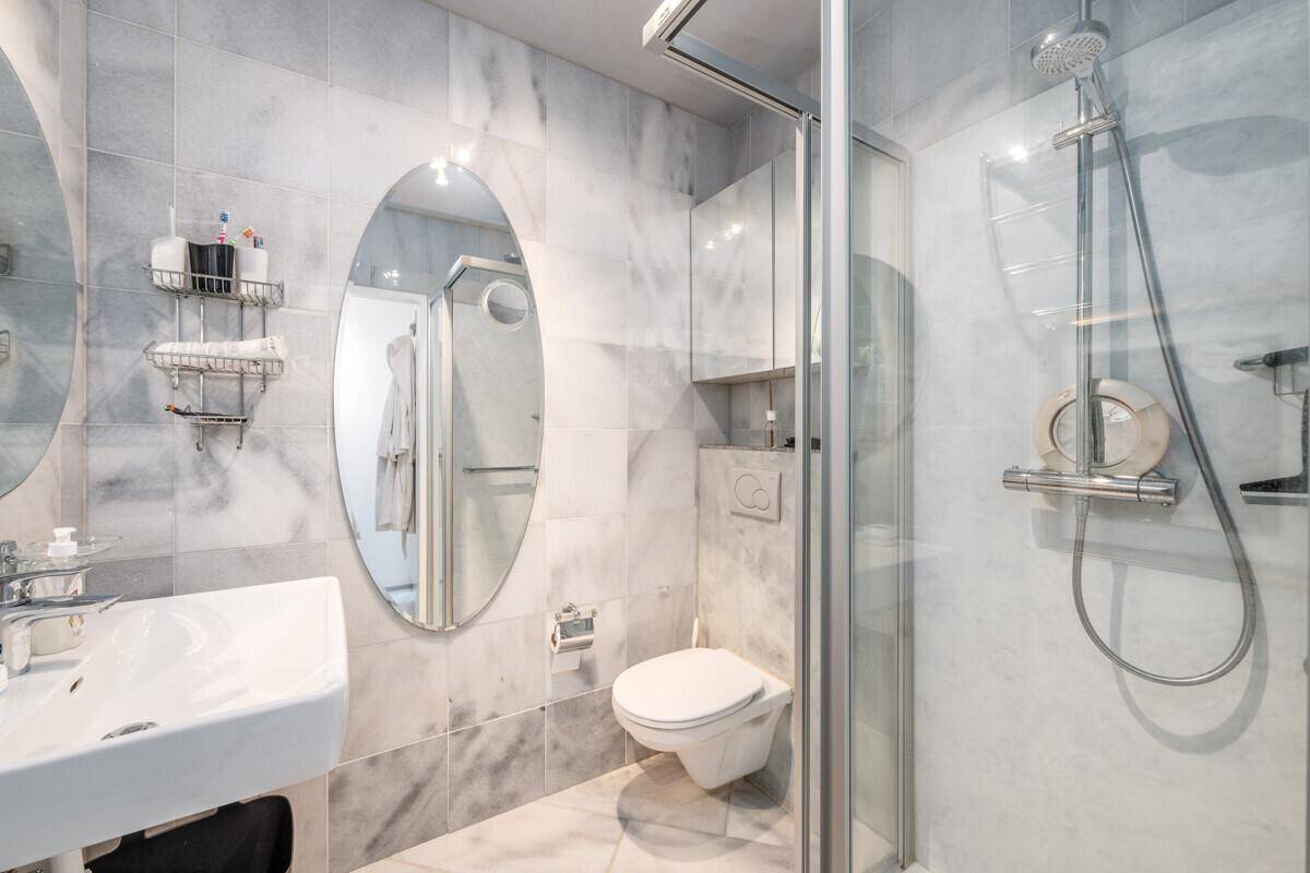 Modernes Badezimmer mit Marmorfliesen, Dusche, WC und ovalem Spiegel über dem Waschbecken.