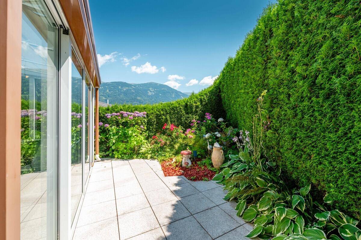 Gepflegter Gartenbereich mit hoher Hecke, Blumenbeeten und Zugang zum Wintergarten.