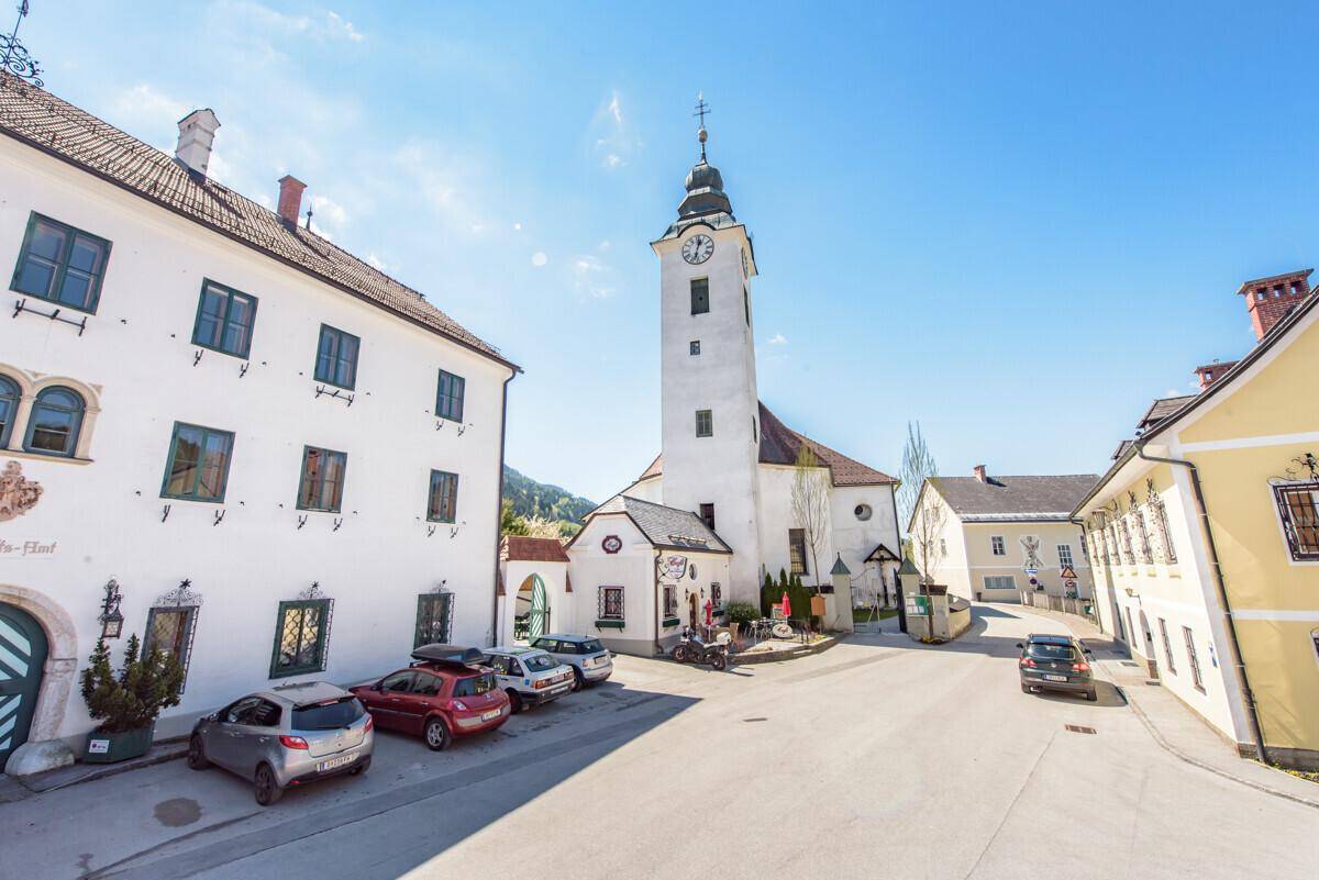 Historischer Dorfplatz mit Kirche, traditionellen Gebäuden und geparkten Autos.