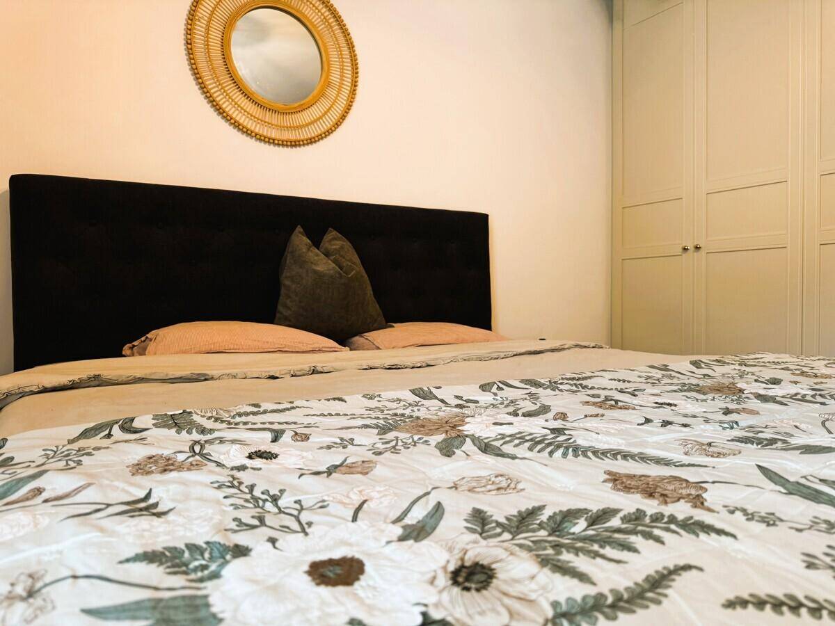 Helles Schlafzimmer mit großem Bett, floraler Bettwäsche und runder Spiegeldekoration.