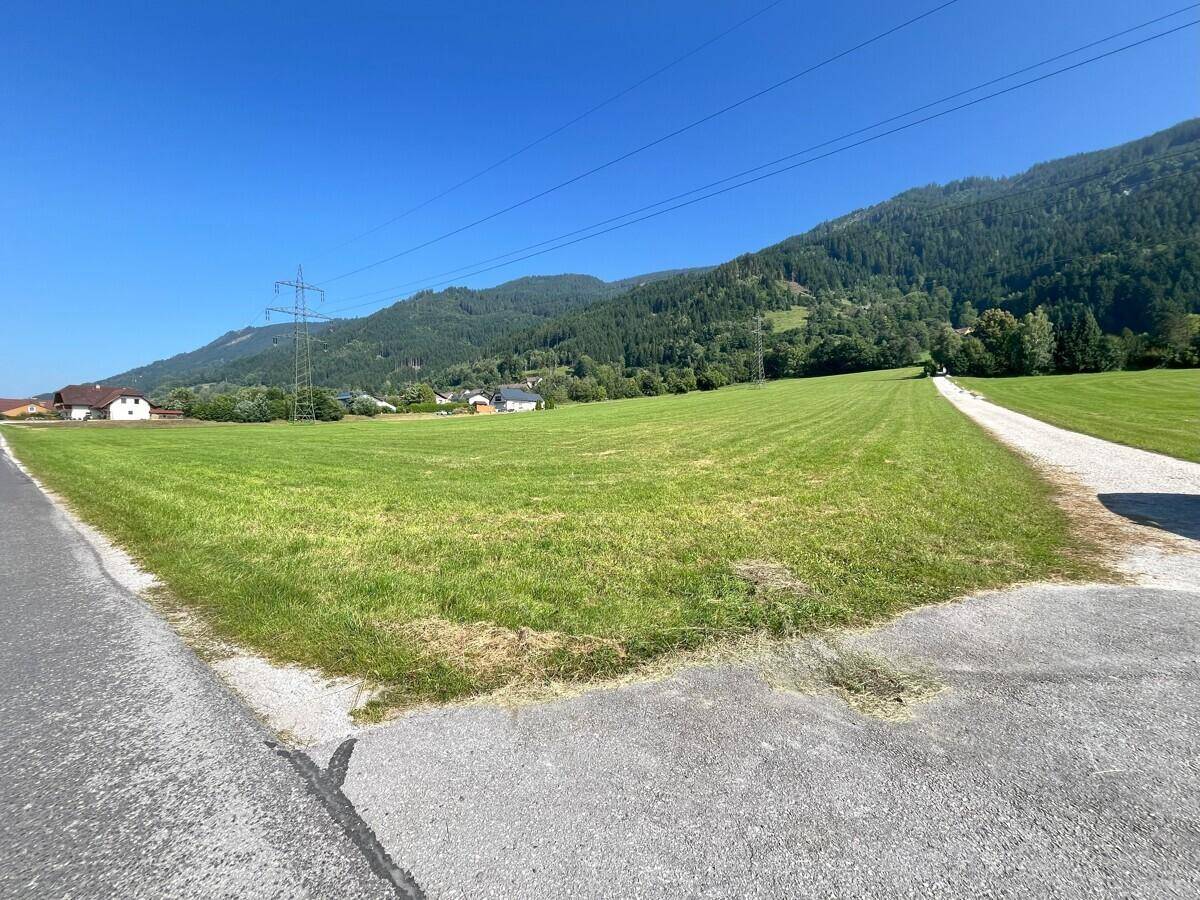 Grünes Feld mit Zufahrtsstraße und Blick auf die ländliche Umgebung mit Bergen im Hintergrund.