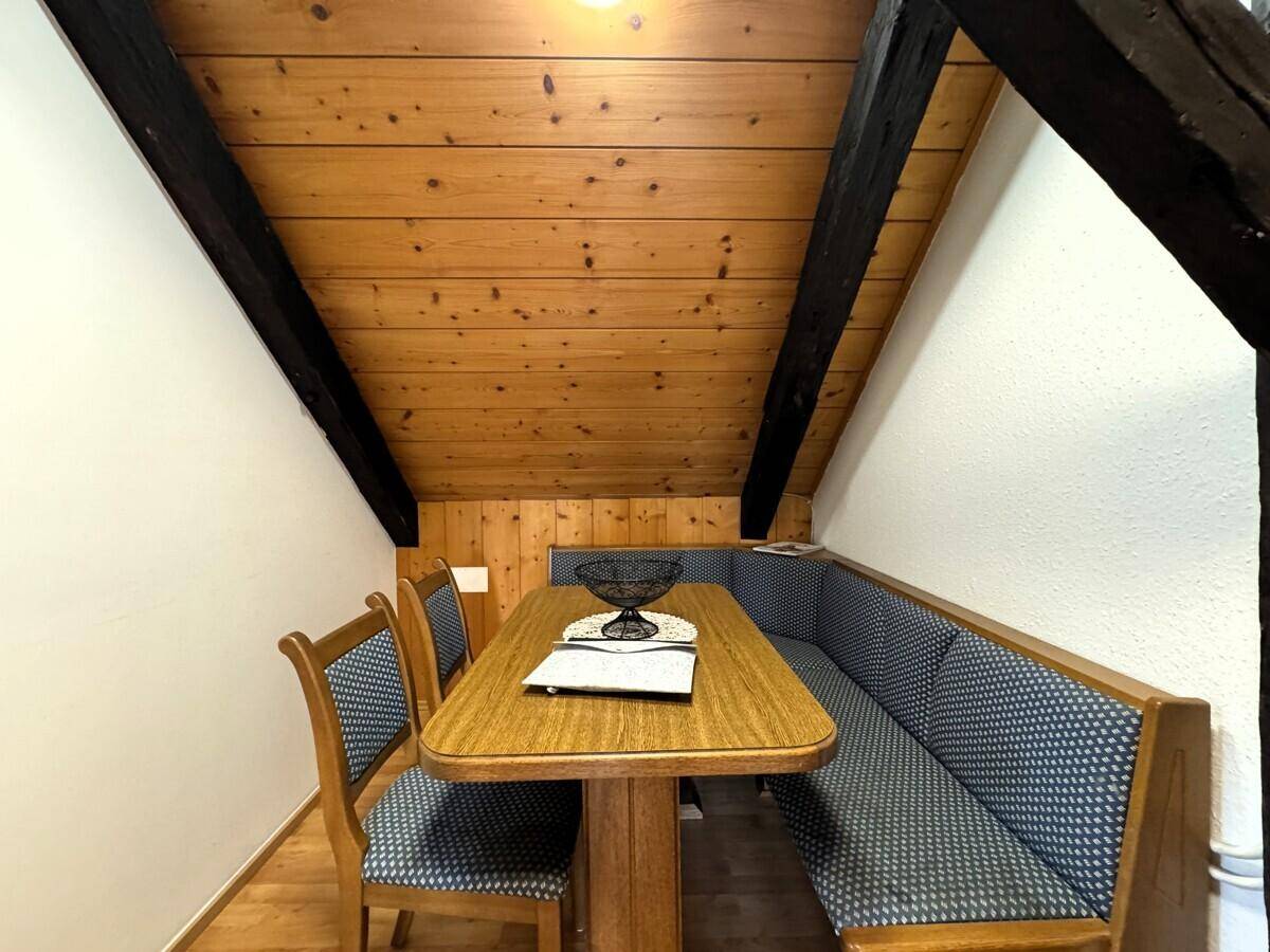 Essbereich mit Holztisch, Eckbank und Holzdachschräge, umgeben von weißen Wänden.