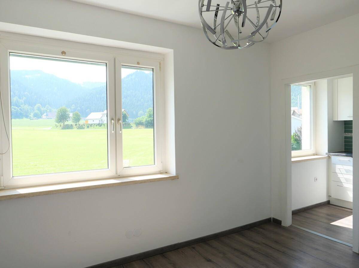 Heller Wohnraum mit großem Fenster, Blick ins Grüne und Zugang zur Küche.