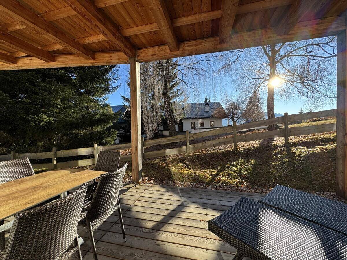 Überdachte Holzterrasse mit Esstisch und Stühlen, umgeben von grüner Landschaft und Bäumen.
