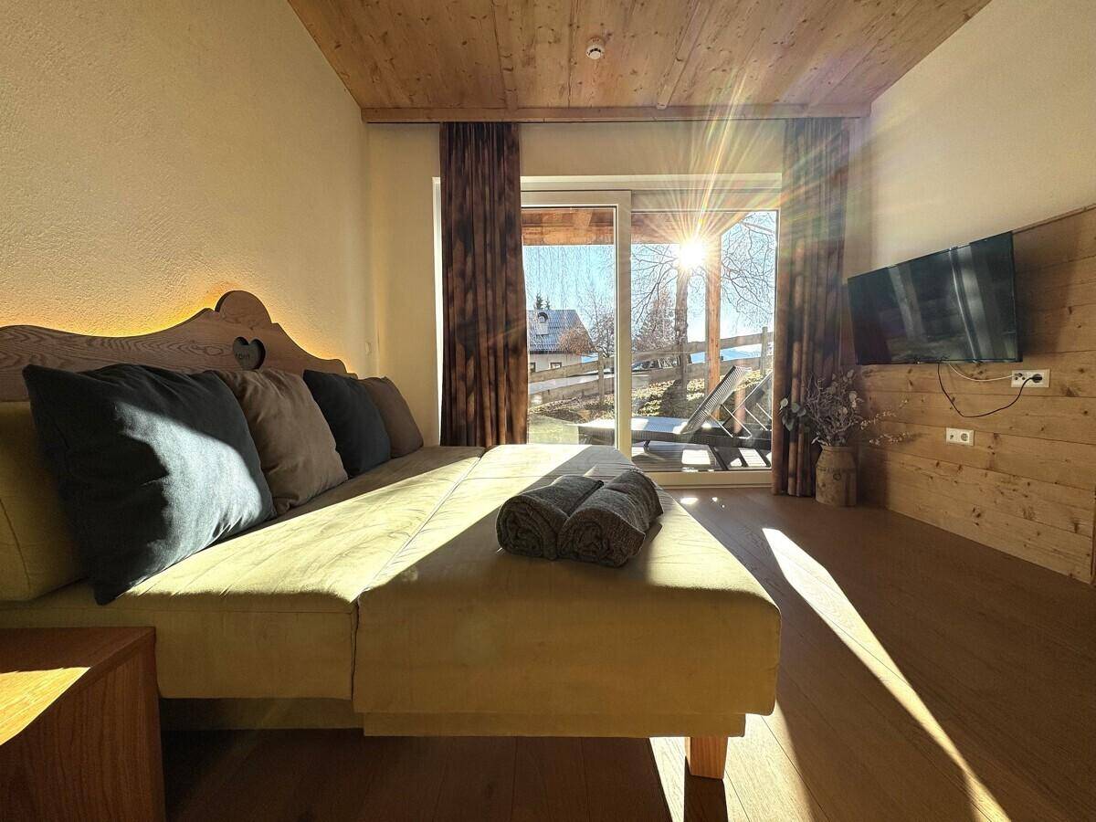 Helles Schlafzimmer mit Doppelbett, Fernseher und großen Fenstern mit viel Sonnenlicht.