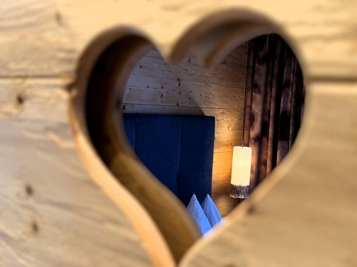 Herzförmiger Ausschnitt in einer Holzwand, der einen Blick in ein gemütliches Schlafzimmer freigibt.