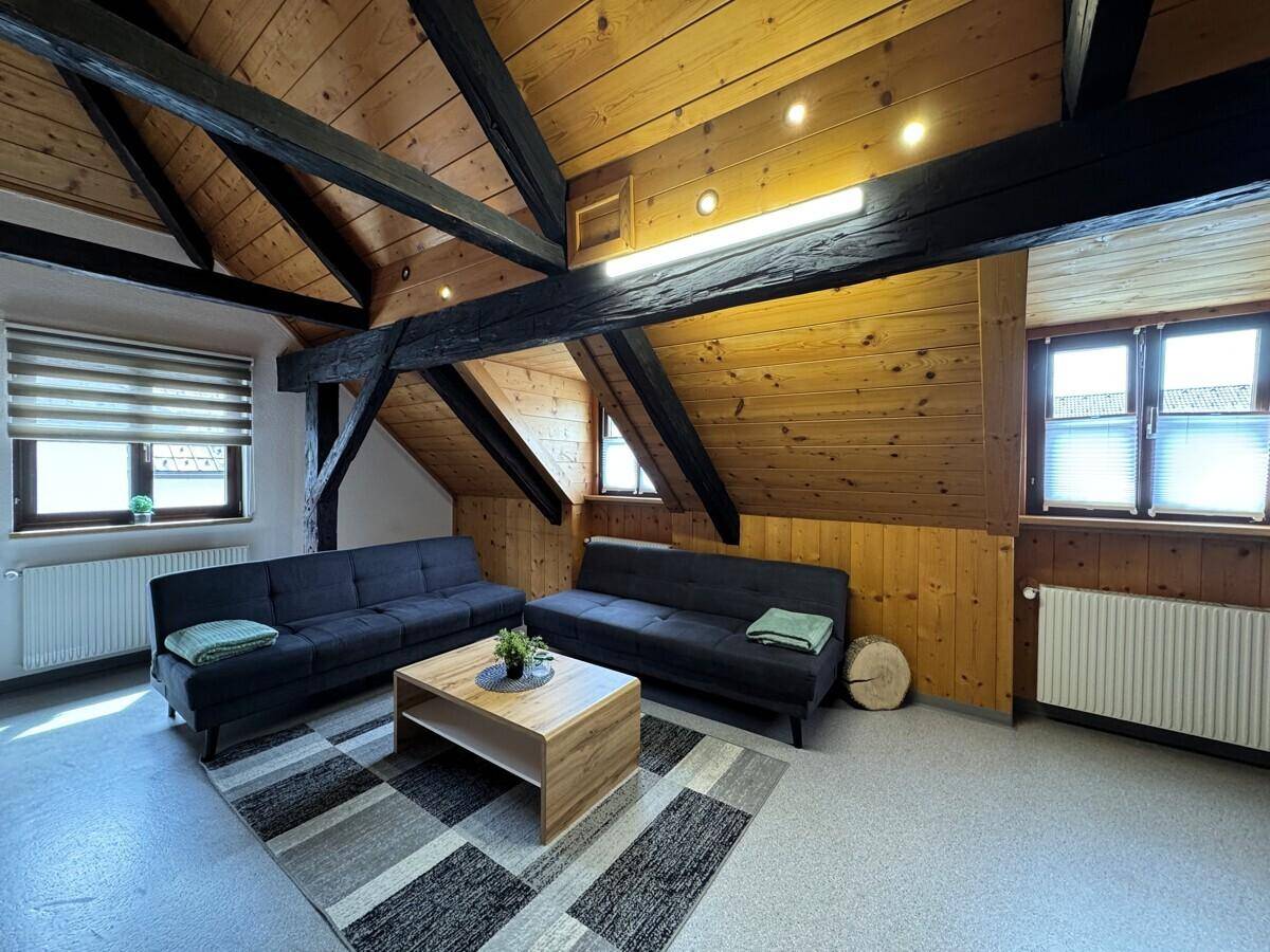 Gemütlicher Wohnbereich unter dem Dach mit Holzbalken und viel Tageslicht durch die Fenster.