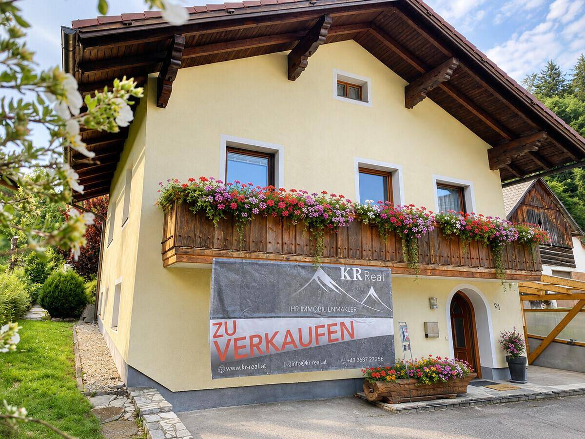 Vorderansicht eines charmanten Hauses mit Blumenkästen am Balkon und einem "Zu Verkaufen"-Schild.