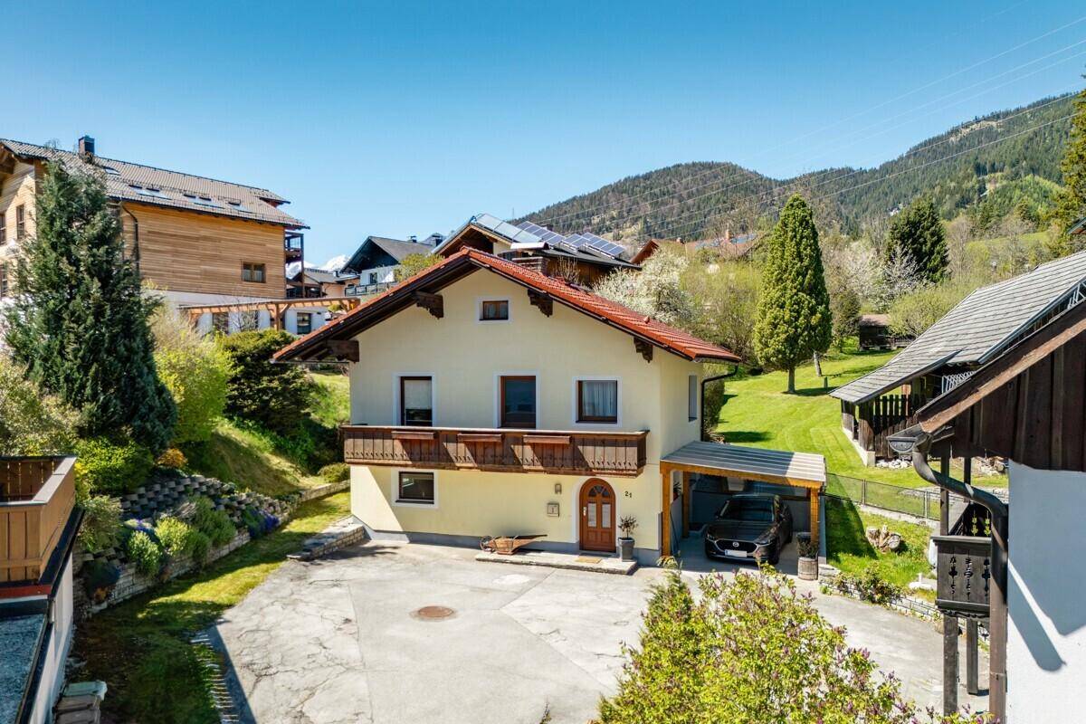 Hausfassade mit Balkon und Carport, umgeben von grüner Landschaft und Bergen im Hintergrund.