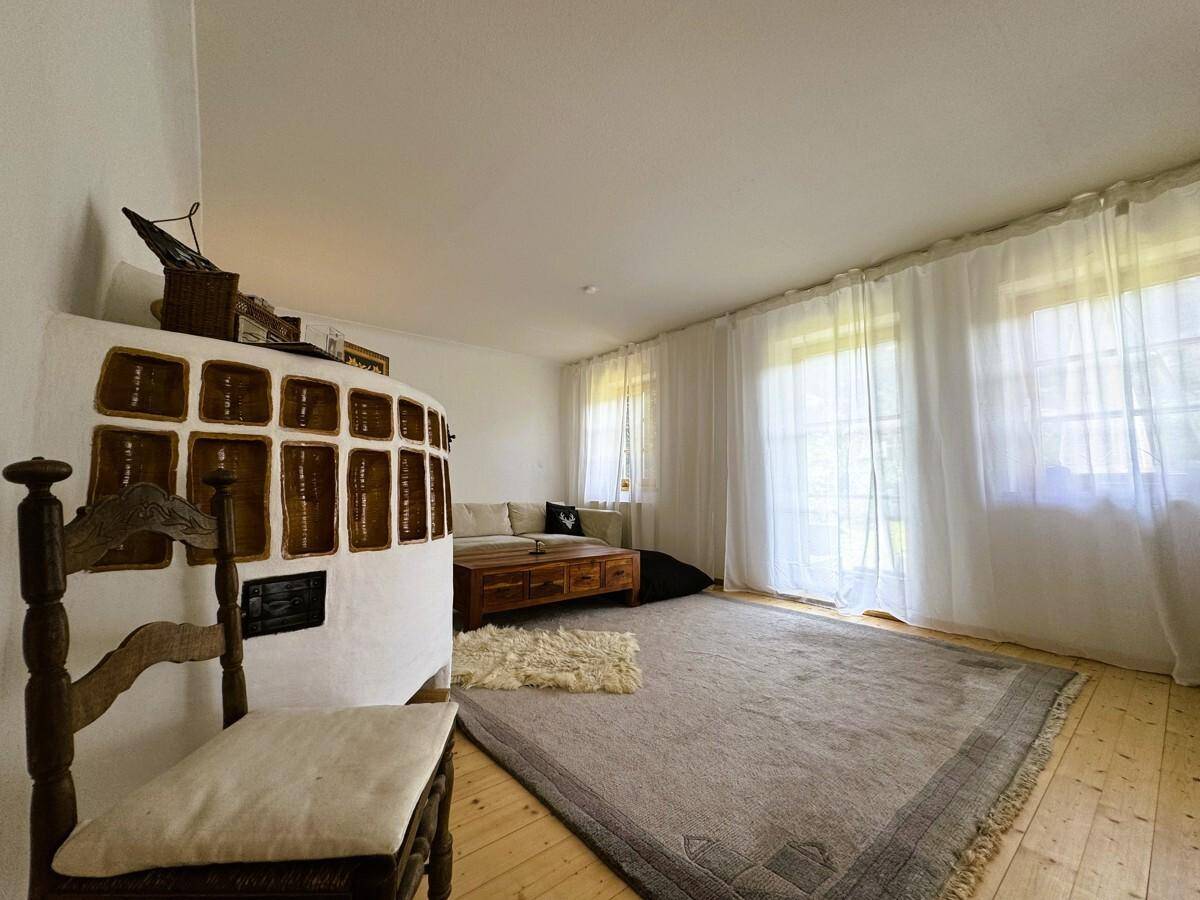 Geräumiges Wohnzimmer mit Kachelofen, hellem Teppich und großen Fenstern.