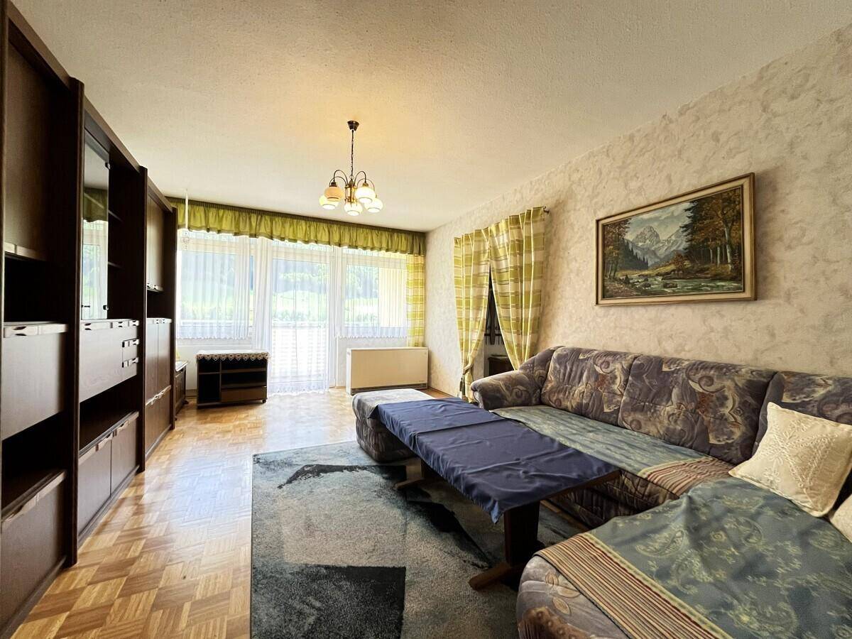 Geräumiges Wohnzimmer mit gemustertem Sofa, großer Holzschrankwand und Parkettboden, hell durch Fenster.