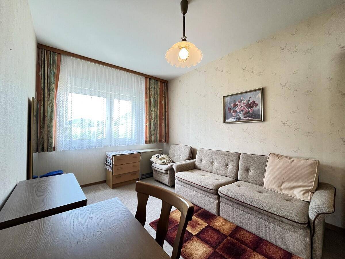 Gemütlicher Wohnraum mit Sofa, Sessel und Holztisch, hell durch das Fenster, mit Teppichboden.