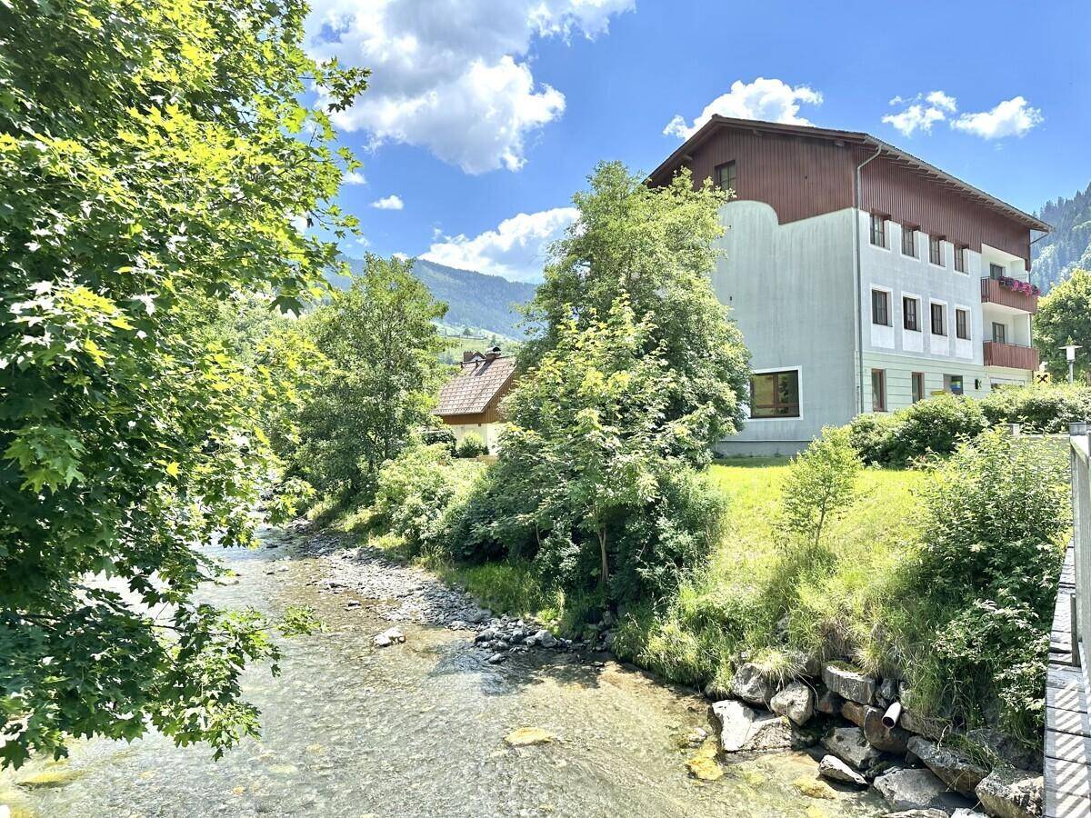 Mehrfamilienhaus am Flussufer mit grüner Umgebung und Bergen im Hintergrund.
