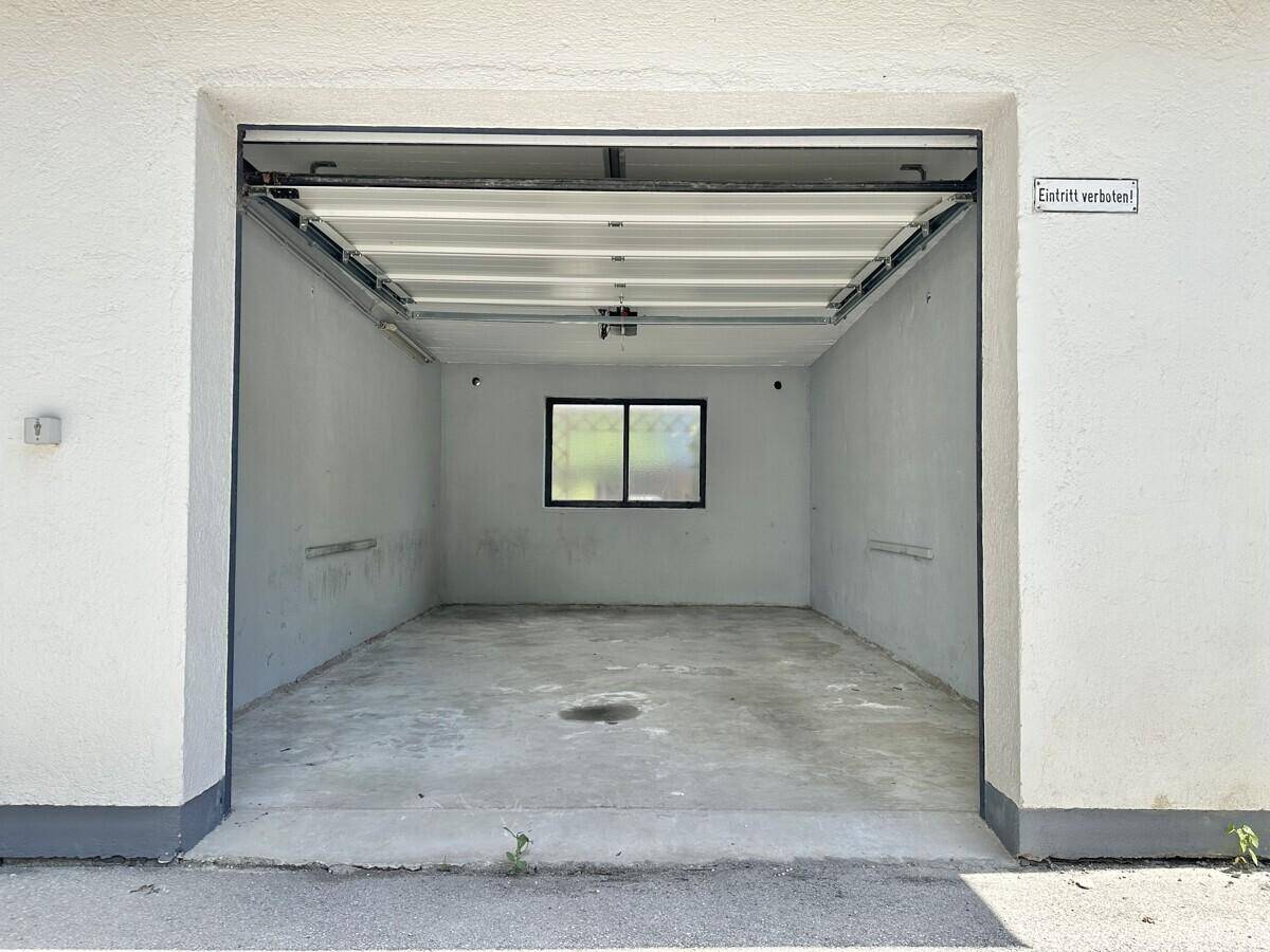 Leere Garage mit Betonboden, einem Fenster und einem offenen Garagentor.