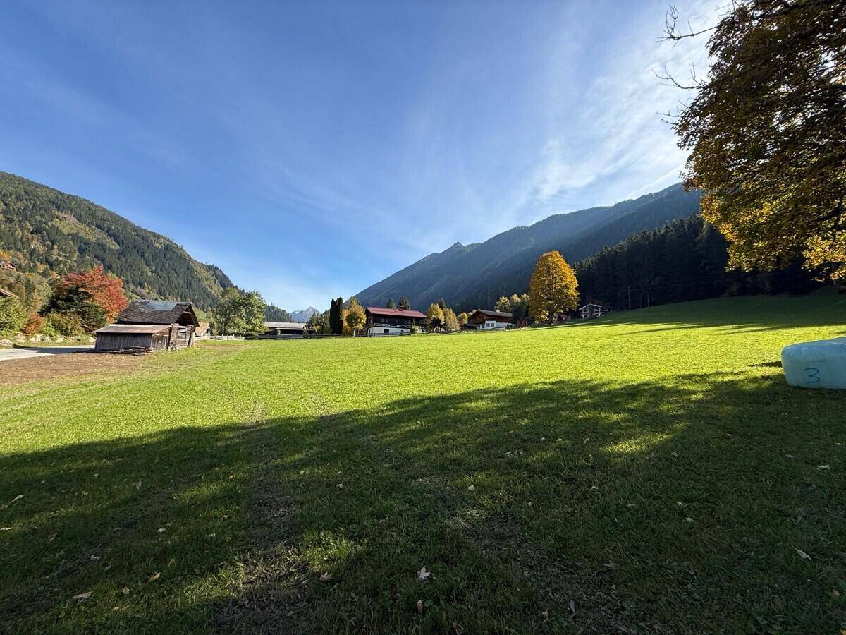 Sonnige Herbstlandschaft mit weiten grünen Feldern, bewaldeten Hügeln und traditionellen Gebäuden in der Ferne.