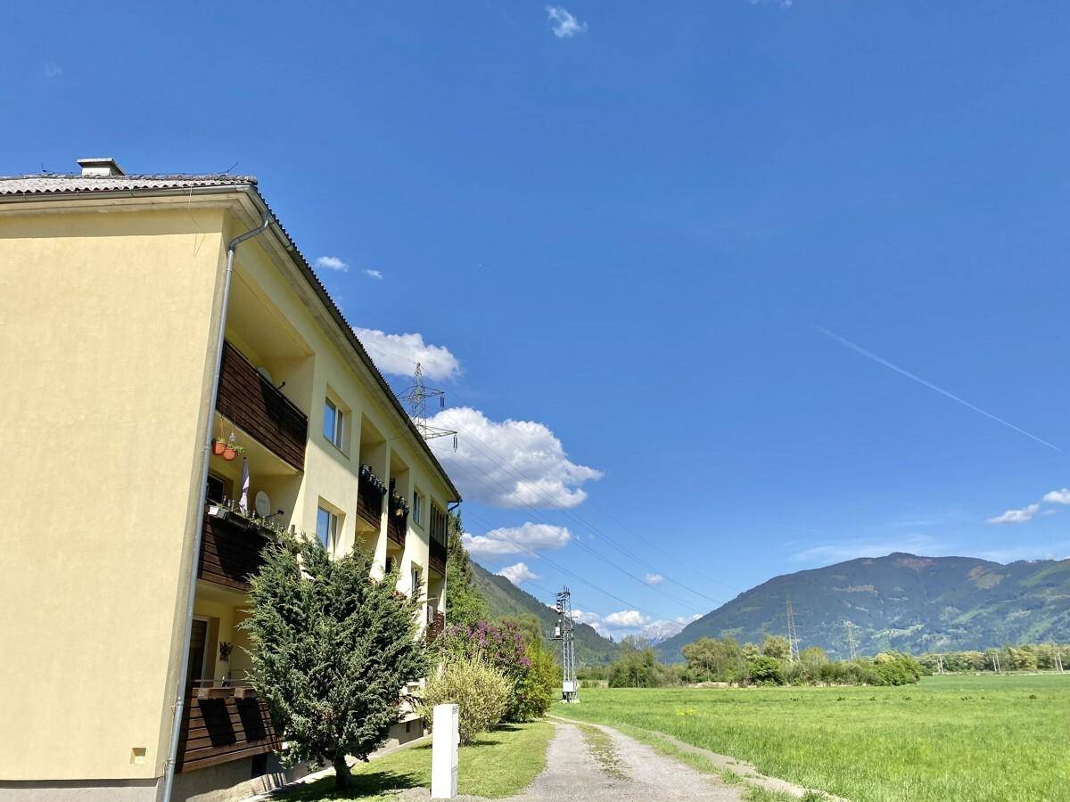 Gelbes Mehrfamilienhaus mit Balkonen neben einer weiten grünen Wiese und Bergen.
